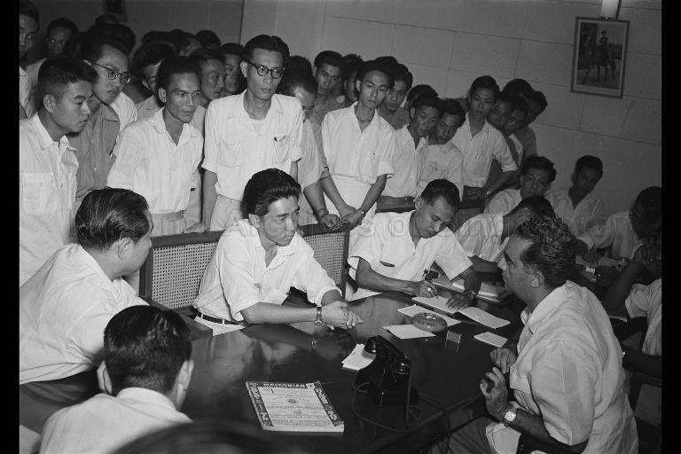 Image shows a group of men sitting and standing around a table. 