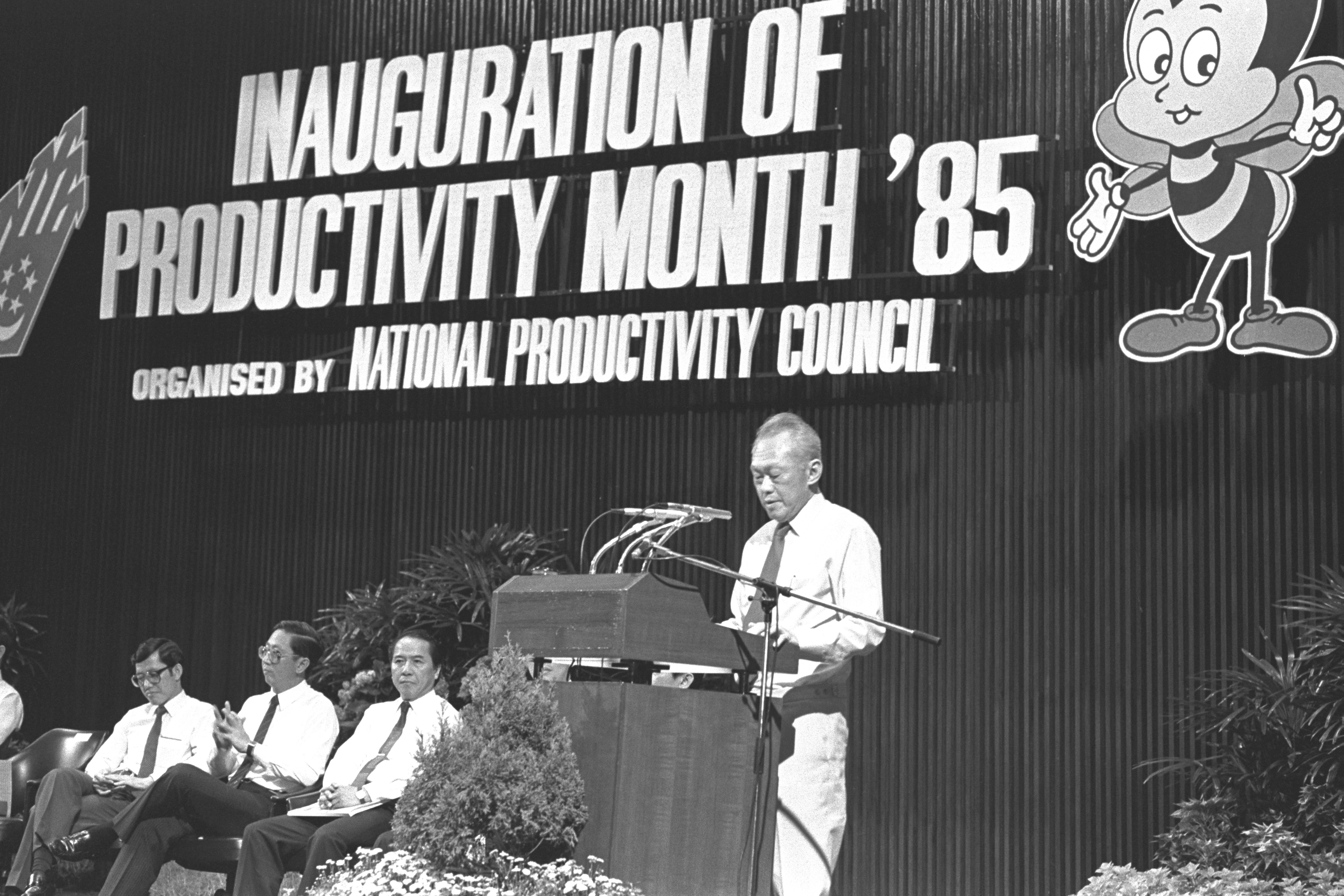 Image shows a man standing at a podium with three other individuals seated to his right.  